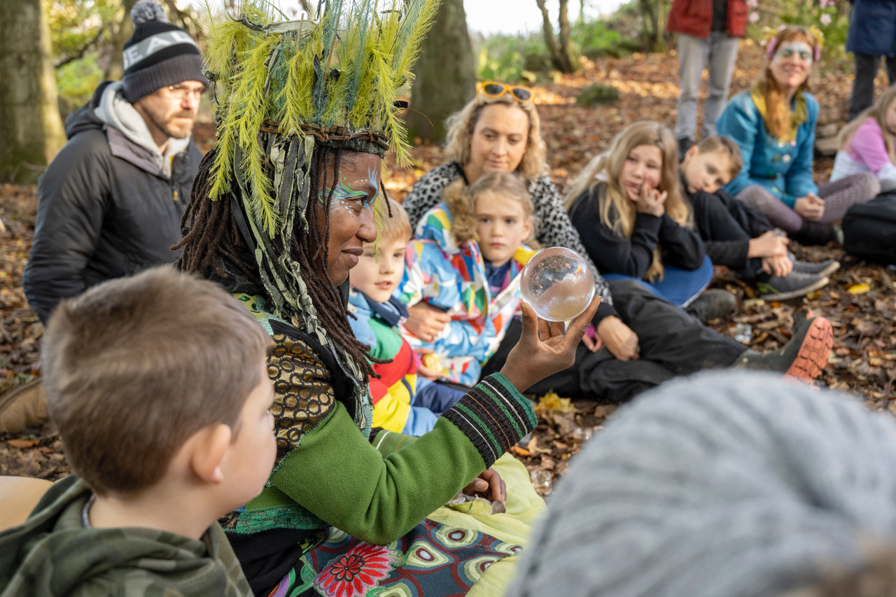 A circus performer holding a glass orb ornament in front of young children and their parents who are sitting down in the woods
